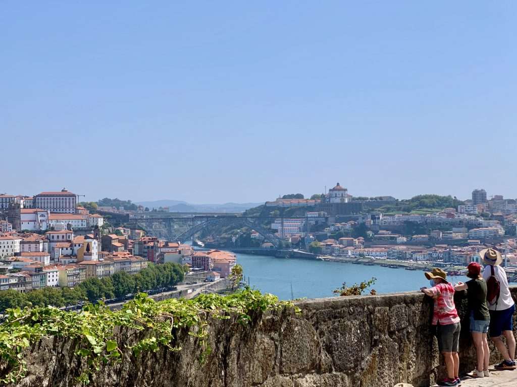 A view from Gaia across the river Duoro toward the Ponte de Luis the 2nd. Three people in shorts and hats look out over a balustrade.