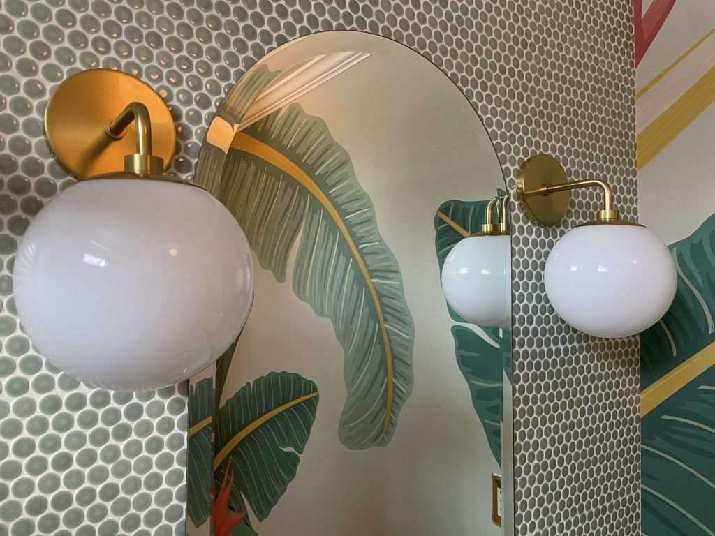 Close up of midcentury brass and white globe wall sconces against green penny tile. A reflection of palm wallpaper is an arched mirror.
